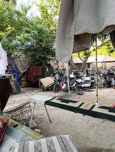 A cluttered outdoor yard in Keŋ-Say, Kyrgyzstan, is depicted in this full-frame, eye-level shot. The ground is composed of dirt and gravel. In the foreground, a patterned rug with dark green and beige geometric shapes is laid out on the ground, partially obscured by a white fabric hanging from the top right corner of the frame. To the left of the rug, a metal table with white legs is set up. Further left, a woven basket rests on the ground next to a cushioned seating area.
The mid-ground of the scene is dominated by several electric scooters and a small, green utility vehicle. One white electric scooter is prominently positioned in the center, surrounded by black scooters. The utility vehicle, covered with a dark red blanket, is parked behind the scooters. A blue wheeled cart is also visible to the left of the utility vehicle. Various cables and electrical cords are strewn across the ground near the scooters.
In the background, a brick wall runs along the left side of the frame, topped by greenery. Trees with dense foliage are present throughout the yard, casting shadows on the ground. Several structures with corrugated metal roofs are visible in the far background. The overall impression is one of a utilitarian or storage space, possibly a yard or workshop, filled with miscellaneous items. No people are visible in the image.