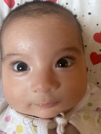 This is a close-up, headshot photograph of a baby. The baby is looking directly at the camera with wide, dark eyes. The lighting is bright, illuminating the baby's face and the white fabric with red heart patterns it is lying on. The background is a soft focus, suggesting a shallow depth of field. The photograph appears to be taken indoors, possibly in a home environment. There is no information to suggest the location is in Mashrab, Uzbekistan.
