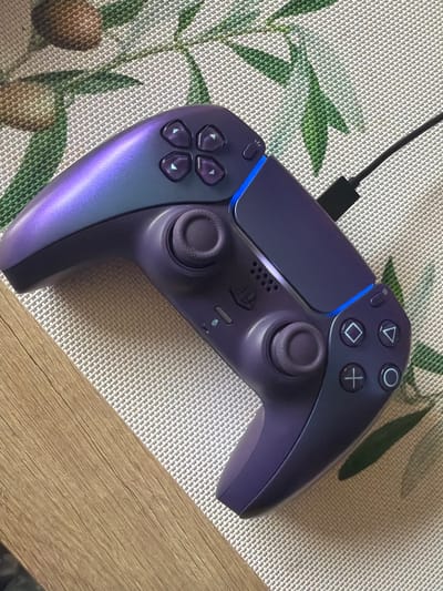 A purple PlayStation 5 Dualsense controller sits diagonally on a patterned placemat, a corner of which rests on a light brown wooden surface. A black charging cable is plugged into the controller. The placemat has a woven texture and is patterned with indistinct green and brown designs. The lighting is soft and diffused. The location is not discernible from the image.