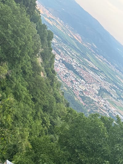 This media file captures a sweeping, high-angle view of a mountainous landscape and a town nestled in a valley. The primary subjects are the natural scenery and the urban development. In the foreground, a dense expanse of lush green trees occupies the lower half of the frame, creating a textured, verdant foreground. These trees appear to be on a steep slope, suggesting a mountainous or hilly terrain.
Beyond the trees, the scene opens up to reveal a town sprawling across the valley floor and up the opposite mountainside. The town is characterized by a dense collection of buildings with red-tiled roofs, interspersed with lighter-colored structures, likely modern buildings. The layout of the town suggests it follows the contours of the land. Agricultural fields or terraced slopes, appearing as green and brown strips, are visible on the mountainsides surrounding the town, indicating a rural or semi-rural setting integrated with the urban area.
In the far distance, a hazy, blue-gray mountain range forms the backdrop, fading into the atmospheric perspective. The sky at the top left of the image is a light, hazy gray, suggesting it might be a cloudy day or that the photo was taken at a time of day when the light is diffused, possibly early morning or late afternoon. There is no discernible text in the image. The overall impression is one of a scenic overview of a town situated in a valley, surrounded by mountains and vegetation. The perspective implies the photo was taken from a higher vantage point, looking down and across the valley. The activity taking place is simply the existence of the town and the landscape itself, with no dynamic events or people prominently featured. The location context of Delchevo, Bulgaria, further places this scene in a Balkan mountain region.