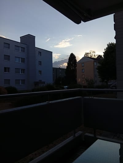 This is an outdoor photograph taken from a balcony in Langenthal, Switzerland, looking out towards apartment buildings and trees at sunset. The sky is a pale blue with some scattered clouds, and the sun is setting behind a cluster of trees, casting a warm glow. In the foreground, a dark railing and a table are visible on the balcony. To the left is a tall, grey apartment building with numerous windows. To the right, a smaller, tan-colored apartment building with a pitched roof is partially obscured by trees. The overall scene depicts a residential area during twilight. There are no people visible in the image.