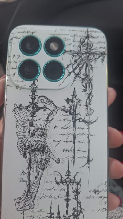 A person's hand is holding a white smartphone with a black and white artistic design. The design features a sketched angel with wings and flowing robes on the left side, and ornate crosses and decorative elements on the right. The background of the phone case appears to be covered in a pattern resembling handwritten script. The phone's camera array is visible in the upper left corner, with four circular lenses. The hand holding the phone has red nail polish. The setting appears to be indoors, possibly a car, given the dark background and the slight blur suggesting movement. There is no visible indication of the location being in Мірскі сельскі Савет, Belarus.