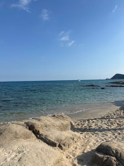 The image depicts a tranquil coastal scene at a beach in Ramatuelle, France. The upper half of the frame is dominated by a clear, light blue sky with sparse, wispy white clouds. A distinct horizon line separates the sky from the sea. The sea exhibits varying shades of blue and turquoise, with clearer, lighter water closer to the shore revealing the sandy bottom. Minimal wave activity is present.
In the mid-ground, a white motorboat is visible on the water, positioned slightly right of the center. To the right, several dark, partially submerged rock formations are scattered in the sea. A distant landmass, appearing as a dark, vegetated hill or cape, is visible on the far right horizon.
The foreground consists of a sandy beach, characterized by light beige sand. Two large, weathered, light brown boulders occupy the lower left and center-bottom of the frame, partially submerged by the encroaching tide. The sand shows numerous footprints, indicating recent pedestrian presence. No individuals or discernible human activity are visible on the beach or immediate vicinity. The scene is brightly illuminated by direct sunlight, casting subtle shadows from the foreground rocks onto the sand.