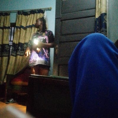 The scene depicts an indoor domestic environment in Umuoji, Nigeria, under low ambient light conditions. In the midground, a standing individual with dark hair, wearing a dark graphic t-shirt with a light-colored design and dark shorts, holds an illuminated rectangular object, consistent with a smartphone screen, directly in front of their torso. The light from this object illuminates their hands and upper body. Their head is tilted slightly back, and their gaze appears directed towards the light source. To the left of the individual, a dark piece of furniture with a red cushion is partially visible. The background features a light blue wall on the left, adorned with two sets of drawn beige curtains featuring dark patterns. To the right of the individual is a dark brown wooden paneled door with a light-colored doorknob. In the foreground, an indistinct mass draped in royal blue fabric occupies the right side of the frame, and a light-colored, out-of-focus object is visible at the bottom left.