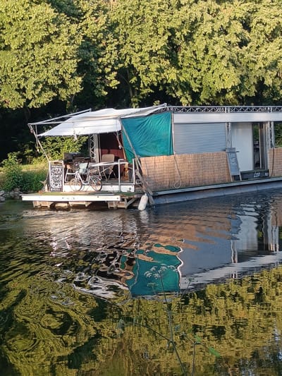 This is a vertical, full shot image of a houseboat on a calm body of water, likely a canal or river, under a bright, sunny sky. The houseboat, which appears to be a commercial establishment with seating and a bar area, is docked alongside a lush green treeline. The structure of the houseboat is visible, with a white awning extending over a deck area. On the deck, a bicycle is parked next to a table and chairs. A blackboard with writing is propped up near the bicycle. The side of the houseboat is adorned with bamboo-like siding, and a teal tarp is draped over a portion of the structure. The water reflects the sky, trees, and parts of the houseboat, creating a rippled, abstract pattern. The location is identified as Haacht, Belgium.