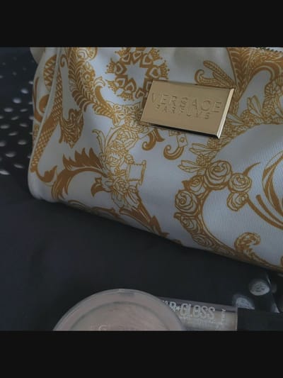 The image shows a close-up of a white and gold patterned cosmetic bag, likely a makeup bag, with the "VERSACE PARFUMS" logo embossed on a gold plaque. The bag is resting on a dark, textured surface, possibly fabric. Below the bag, a portion of makeup product, appearing to be a pressed powder compact and a lip gloss tube, is visible. The lip gloss tube has "STAR GLOSS" written on it. The lighting is soft, highlighting the texture of the bag's pattern and the metallic sheen of the logo and lip gloss. The scene is static and appears to be set indoors. The context of Bielawska Wieś, Poland is not evident from the image itself.
