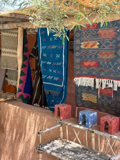 This vertically oriented image captures a vibrant display of traditional textiles and crafts in Tahannaout, Morocco. Numerous intricately patterned rugs and runners are prominently featured, hung from wooden beams. The uppermost rug is dark blue with elaborate geometric designs in red, brown, and cream, and has white fringed edges. Below it, a bright blue runner with smaller, colorful motifs hangs. Further down, various other textiles are visible, showcasing diverse patterns and colors including green, pink, black, and beige.
On the left side of the frame, a thriving olive tree with slender green leaves and wooden branches partially obscures the view, adding a natural element to the scene. To the right, three cubic, perforated stools are arranged next to a bamboo ladder or scaffolding. Two stools are red and one is blue, all appearing weathered with decorative cut-outs. The ground or a wall on the right is a rough, reddish-brown earthen surface, likely made of rammed earth or mudbrick, with visible straw fibers.
The scene is bathed in bright sunlight, indicated by the clear illumination and the shadows cast by the bamboo ladder on the earthen surface, suggesting a sunny daytime. The overall impression is one of a lively, traditional outdoor setting, possibly a market or workshop, highlighting local Moroccan craftsmanship and natural materials. No visible text is present in the image.