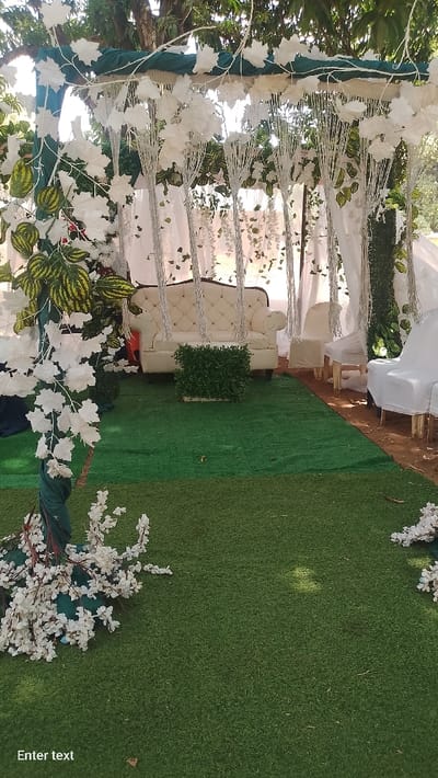 An outdoor event setup in Rinze, Nigeria, features a decorated canopy or arbor. A cream-colored, tufted sofa with dark legs is centrally placed on artificial green turf, flanked by two white fabric-covered chairs on the right. A rectangular planter box containing green artificial shrubs sits in front of the sofa. The overhead structure is draped with dark green fabric and extensively adorned with artificial white leaves, small white flower clusters, and green variegated foliage. Multiple strands of white beads or lights hang vertically from the structure. White sheer curtains are visible in the background, partially obscuring natural trees and bright sunlight. A dark green fabric is wrapped around a vertical support on the left, with white artificial flowers scattered at its base on the turf. No individuals are present in the scene.