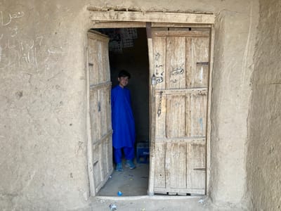 The image depicts a young boy standing in a doorway, partially obscured by the wooden door. He is wearing a blue long-sleeved shirt and pants. The doorway leads into a room with an unidentifiable object on the floor. The doorway is framed by a mud-brick wall, which has several scribblings on it. The scene is located at coordinates 30.532 latitude and 66.909 longitude in the city of Saranan, Pakistan.