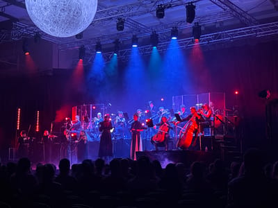 A concert stage is illuminated with a mix of blue, red, and green stage lighting, and an artificial moon hangs overhead. An orchestra performs on the stage, comprised of string and wind instruments. The conductor is in the center. In the foreground, the backs of audience members fill the seats. The location is in Colchester, United Kingdom.