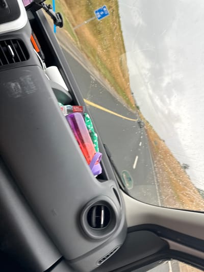 The image depicts the interior of a vehicle, with a focus on the dashboard and the view through the windshield. The dashboard is black and grey, featuring an air vent and a textured surface. On the dashboard, a red and purple bottle is visible, along with a roll of white material and a bright orange object. The windshield shows a road with a yellow line, indicating a highway, and sparse traffic in the distance. The weather appears overcast with rain streaks on the windshield. The landscape outside is characterized by dry grass and a cloudy sky. There is a blue road sign with white symbols visible in the distance. Based on the context of Tubberneering, Ireland, this scene is likely captured while driving on a road in or around that location.