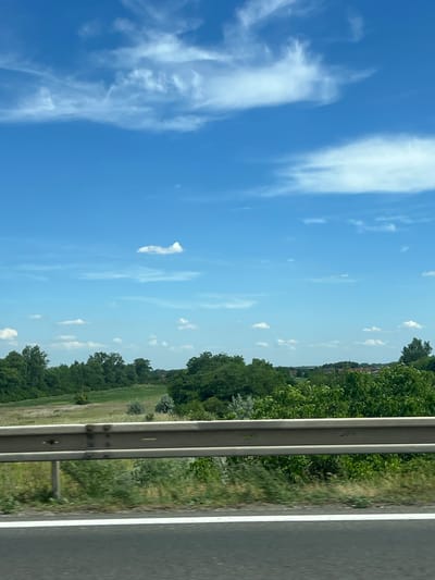 The media file depicts a roadside scene, likely captured while traveling along a highway. The image is oriented vertically, suggesting it might have been taken from a car window.
The primary subjects are the sky, a guardrail, and a landscape. The sky is a vibrant blue, with scattered white clouds, indicating fair weather. The guardrail, made of metal, runs vertically along the right side of the frame, separating the road from the surrounding environment. Beyond the guardrail, a green landscape unfolds, characterized by lush trees, fields, and what appear to be buildings in the distance. The presence of a paved road with a white line marking further confirms the highway setting.
The image captures a daytime scene, with bright lighting and clear visibility. The overall mood is peaceful and serene, evoking a sense of travel and observation of the natural environment. No people are visible in the image, and there is no visible text. The location context of Veľký Biel, Slovakia, suggests this is a scene from that region.