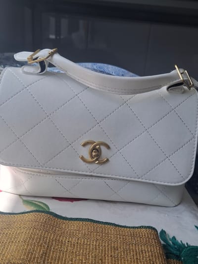 A white quilted handbag with gold hardware is displayed on a bed with floral patterned sheets. The handbag has a flap closure with a gold interlocking "C" logo. A white strap with gold buckles is attached to the bag. To the right of the handbag, a textured gold fabric with green trim is visible, possibly part of a cushion or throw. The scene is lit with artificial light, suggesting an indoor setting, likely a bedroom. The overall impression is one of luxury and style. There is no visible text in the image.