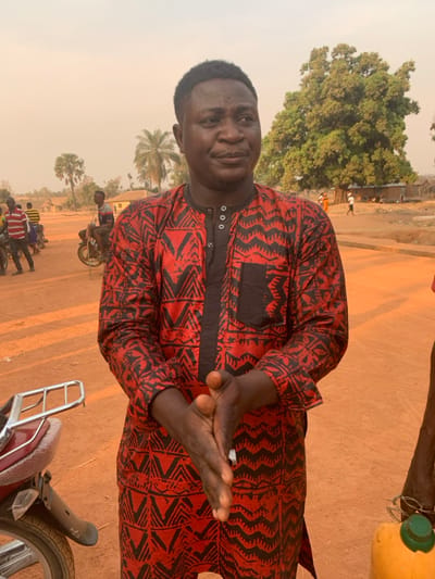An African man stands facing the camera, wearing a red and black patterned shirt. He holds his hands up in a gesture. A green tree is visible in the background. The scene takes place outside, where multiple motorcycles are parked. Other people are gathered nearby. The lighting suggests it might be early morning or late afternoon.