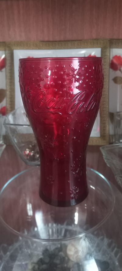 A ruby red glass with the Coca-Cola logo embossed on it sits on a clear glass plate, which is on top of a larger clear glass bowl. The glass has a textured surface with small raised circles and embossed snowflake designs. The background consists of a reddish-brown textured wall above a framed picture with floral motifs. The scene appears to be indoors, possibly in a home setting. There are no people visible in the image.
