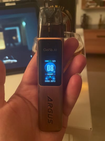 The image shows a close-up of a person's hand holding a vape device. The vape device has a digital screen displaying "Gene AI," a battery indicator at 52%, a wattage of 8W, and "SMART." Below this, there are icons labeled "R" and "P" with corresponding numbers. The device itself is rose gold colored and has "ARGUS" written vertically on its side. In the background, a tablet screen is visible, showing a blurred image. To the right of the vape, a glass with a beverage is partially visible, along with shelves in the background. The setting appears to be indoors, possibly a home or a bar, with warm lighting. There is no specific indication of the location being in Yunus Emre, Turkey.