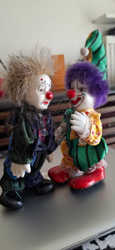 This media file features two porcelain clown dolls lying on a reflective, gray surface. The dolls are positioned on their sides, facing different directions.
The top doll has a bright yellow outfit with red polka dots and white lace trim. It wears a green and black striped hat with a purple, fuzzy wig. The clown's face is painted with a red nose, drawn-on tears, and a smile. Its hands are white porcelain.
The bottom doll has a more muted color palette. Its hair is a light brown, wispy material. It wears a dark blue and green outfit with a black net collar. The clown's face also has a red nose, painted tears, and a smiling expression with rosy cheeks. This doll's clothing appears layered and somewhat disheveled.
The dolls are lying on what appears to be a desk or table, with the reflective surface catching light. In the background, there are out-of-focus elements, including what looks like blinds or a radiator and possibly furniture. The lighting suggests an indoor setting during the daytime. There is no visible text in the image. The overall scene evokes a slightly eerie or melancholic mood, common with depictions of dolls.