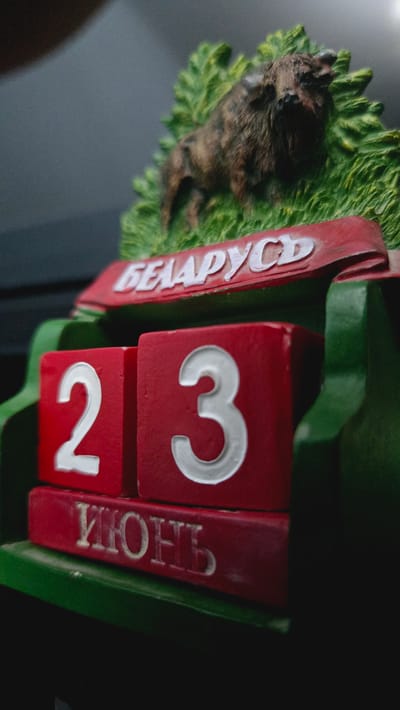A close-up, low-angle shot of a red and green wooden perpetual calendar is shown. The calendar displays the date "23" in large white numbers on red blocks. Below the numbers, the word "ИЮНЬ" (June in Cyrillic) is carved into a red block. A red banner above the date blocks reads "БЕЛАРУСЬ" (Belarus in Cyrillic) in white lettering. A miniature brown bison figurine stands on a green grassy base above the banner. The background is dark and out of focus. There is no indication of Kaka, Turkmenistan in the image.