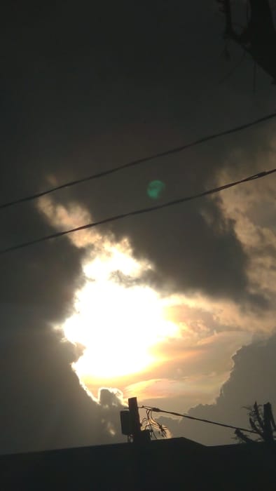 The image displays an outdoor scene over Longuo 'A', Tanzania, characterized by a heavily clouded sky during either sunrise or sunset. A powerful, brilliant white and yellow light source, consistent with the sun, radiates through a large irregular opening in dense, dark grey and black clouds positioned centrally in the frame. The edges of the clouds immediately surrounding this aperture glow with bright yellow and orange hues. Two dark, horizontal lines, identified as overhead power or communication cables, are visible in the upper-middle portion of the sky, spanning across the cloud formations. A distinct green circular lens flare is present in the upper sky, slightly to the right of the image's vertical center. The lower portion of the image features a dark, silhouetted rooftop or structure. From this silhouette, a vertical utility pole or antenna mast, accompanied by multiple entangled wires, rises slightly to the left of the frame's center. Another smaller, dark, silhouetted object, possibly a damaged tree branch or additional utility component, is visible on the far right. No individuals or discernible actions are present within the scene.