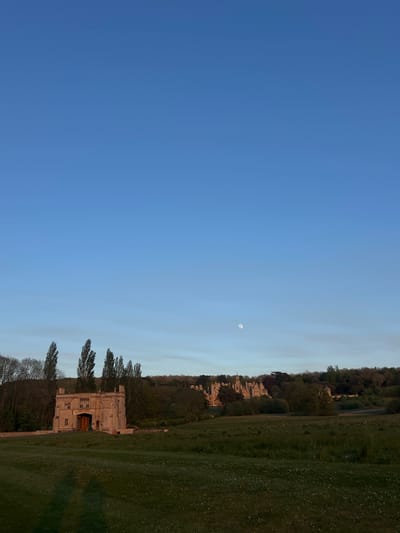 A panoramic outdoor scene captures a tranquil landscape under a clear blue sky. The image, oriented as if viewed in landscape mode, features a vast expanse of bright blue sky occupying the upper two-thirds of the frame. In the mid-right portion of the sky, a distinct, pale moon is visible, suggesting either early morning or late afternoon/dusk.
The lower third of the image is dominated by a wide, slightly uneven grassy field, dotted with lighter patches that could be wildflowers or dry grass. Beyond the field, a line of tall, slender trees stands in front of a substantial, light-colored stone building. This structure, possibly a historic manor house or estate, features multiple stories, numerous windows, and intricate architectural details. A grand entrance with large wooden doors is visible on one side of the building. Behind the building and further into the background, a dense line of darker trees forms a natural backdrop, blending with the distant horizon.
No people or specific activities are discernible, contributing to the peaceful and serene atmosphere. The weather appears clear and calm, with no visible clouds. The architecture and landscape are consistent with the location context of South Kesteven, United Kingdom, evoking a sense of traditional English countryside and historic estates. There is no visible text in the image.