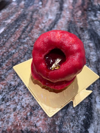 A close-up, high-angle, vertical shot focuses on a red, donut-shaped dessert with a glossy finish and small water droplets, resting on a gold paper doily. The dessert has a hole in the center, revealing a dark, glossy filling and a piece of gold leaf. The doily is square-shaped with a notched corner. The dessert and doily are placed on a speckled granite countertop with shades of gray, black, white, and pink. The image was taken in Wisconsin Dells, United States of America.