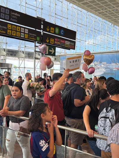 The image depicts a crowded indoor waiting area, likely an airport terminal, given the signage for "Lavabos," "Toilets," "Aseos," and various transportation services like "Metro," "Taxi," "Uber," "Cabify," "Bolt," "Llançadora Bus-shuttle," and car rental companies such as "Avis," "Goldcar," and "Europcar." The location is identified as El Prat de Llobregat, Spain, based on the airport context.

In the foreground, a young girl wearing a blue soccer jersey with a UEFA Champions League patch is visible, looking to her right with her hands near her face. To her left, a woman in a dark grey patterned top and ripped white jeans is leaning on a metal railing. Behind her, a man in a white shirt holds a bouquet of pink and white flowers.

Further back, several people are gathered, some looking towards the entrance or waiting. There are multiple directional signs suspended from the ceiling. A cluster of balloons, including a large pink heart-shaped one and two smaller round balloons (one pink, one metallic gold), are floating near a sign. Another sign, held by a person in the middle ground, features text in Korean characters.

The environment appears to be a modern building with large glass windows and a metallic ceiling structure. The overall atmosphere suggests an arrival or departure area where people are waiting to greet or meet others.