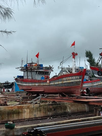 The scene depicts a shipyard or dry dock facility in Thuận An Ward, Vietnam, under an overcast sky. The foreground features stacks of rusted metal beams on a dirt and gravel ground, adjacent to a concrete ledge holding a green plastic basin and a white container. In the midground, several wooden fishing boats are elevated on dry land, supported by wooden blocks and metal scaffolding or rails.

The central vessel, a large red-hulled fishing boat, has "TTN 90612 TS" painted in white on its bow. Its hull also features a dark gray stripe and a narrower light gray stripe. The boat's superstructure includes a cabin, masts with rigging, and several blue barrels on its upper deck. Two red flags, consistent with the flag of Vietnam, are flown from masts on the bow and stern sections of this boat.

To the left, another red and gray fishing boat is partially visible, also elevated. To the right, a third boat, primarily red with some white and blue, is also docked. The immediate ground around the boats consists of dirt, concrete structures, and embedded metal tracks. In the distant background, a small blue building or structure is partially visible, with indistinct human figures near it, suggesting ongoing activity. Scattered evergreen trees are visible behind the boats, and numerous small birds are visible in the pale gray sky. No explicit human actions or interactions are discernible in the immediate vicinity of the primary boat.