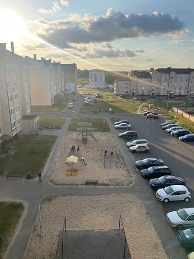 The image captures a view of an apartment complex courtyard in Malaryta, Belarus, during what appears to be sunset or sunrise, indicated by the low angle of the sun and the soft, warm lighting. In the foreground, a sand-filled playground with swings and a sandbox is visible, with a few children present. To the left, a paved pathway leads to a grassy area with benches and a trash can. Adjacent to the playground is a parking lot filled with various cars. The apartment buildings, with their light-colored facades and multi-story windows, dominate the background, with trees and a distant building visible beyond. The sky is partly cloudy, with the sun's rays piercing through.