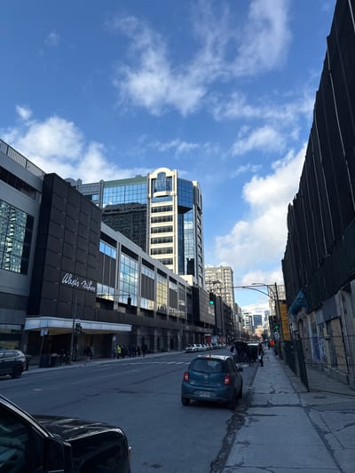 This image shows a street view in Westmount, Canada, with buildings lining both sides. The sky is blue with scattered clouds. On the right side of the street, a blue car is in motion, with several parked cars visible along the curb. Pedestrians are seen gathered at the building entrance marked with "Alexis Nihon". Above them, multiple high-rise buildings are visible, reflecting the sky. Construction scaffolding or a dark overhang is visible above the left side of the street.