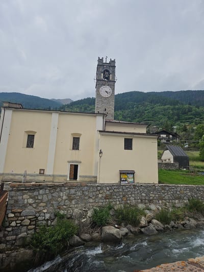 A stone clock tower, part of a pale yellow church building, rises against a cloudy sky and a backdrop of green, tree-covered mountains. The clock face, visible on the tower, shows the time as approximately 10:10. The church has two arched windows with decorative metalwork, and a single entrance with an open wooden door. A stone wall, constructed from various sized rocks, runs in front of the church and alongside a fast-flowing river. The riverbed is filled with large stones and some green vegetation. Beyond the church, houses are nestled on the hillside. The image appears to be taken from across the river, looking towards the church and clock tower in the town of Ponte di Legno, Italy.