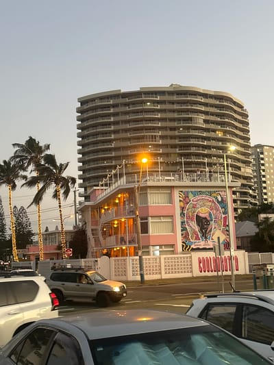 This image depicts an evening street scene in Coolangatta, Australia, showcasing a dynamic interplay of modern architecture, vibrant art, and ambient lighting.
On the left, a tall, multi-story building stands prominently with a distinctive, curvilinear design. Its facade is characterized by stacked, wavy balconies that create a fluid, undulating pattern, with lights visible from inside numerous windows.
Adjacent to this large structure, on the right, is a smaller, pink-colored building. A significant feature of this building is a large, colorful mural on its upper exterior. The mural displays intricate patterns of leaves and abstract shapes, framing a dark, stylized animal, possibly a panther, within a large 'C' shape. Text fragments like "EXTRAORDINARY" and "COOLANGATTA" are visible within or near the mural. Below the mural, the pink building features windows and is adorned with festive string lights. A prominent sign with red lettering spelling "COOLANGATTA" is affixed to a white wall next to the mural. A faint sign reading "PINK HOTEL" is also visible further down.
In the foreground and to the right, several vehicles, including a white SUV/van and other cars, are present on a paved street. The street is lined with palm trees, which are festooned with draped string lights, contributing to a festive atmosphere. Streetlights and building lights are illuminated, indicating the time of day as dusk or early evening, with the sky appearing a pale grey. A partially obscured red "DO NOT ENTER" sign is visible on a post near the road. The overall scene suggests a lively coastal environment as day transitions to night.