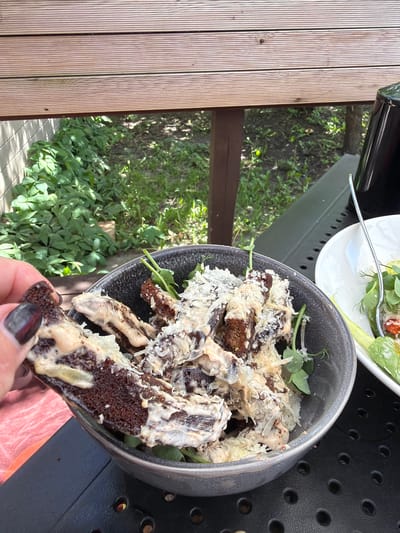 The image captures a close-up, high-angle shot of a salad being held by a hand with dark-polished nails. The salad appears to be a mix of greens, dark bread croutons, and a creamy dressing, possibly garnished with shredded cheese or a similar topping. The salad is in a dark-colored bowl. A second plate with what appears to be a salad with light-colored vegetables and dill can be seen in the background. The table beneath the bowl is black with a perforated design. The background includes a wooden deck and greenery, with a glimpse of what might be a garden. The location is likely Ragaciems, Latvia, considering the context provided. No other people are visible in the frame.