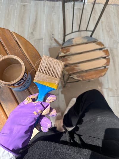 Here's a detailed description of the image:
The image shows a first-person view of someone painting outdoor furniture. The location is in the city of Береговой сельский совет, Ukraine.
The person is wearing purple rubber gloves with paint splatters and is holding a paintbrush with a blue handle, yellow insert, and tan bristles. The bristles are coated in tan paint. The paintbrush is positioned above an open can of tan paint. The paint can has Cyrillic text on the label.
Below the paint can is a small, round, wooden table. The table is made of wooden slats that are painted tan. The table is sitting on a tiled surface with a wood-grain pattern.
Behind the table, a chair with a metal frame and wooden slats on the seat is partially painted tan.
To the right, the person is wearing black clothing and white shoes. The entire scene is lit by natural sunlight.