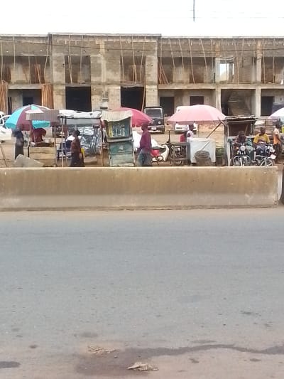 The image depicts a street scene in Ihiala, Nigeria. A wide concrete barrier divides the foreground road from the midground activity. Behind the barrier, vendors have set up stalls under colorful umbrellas, selling goods. Several people are visible, including vendors and customers. There are also motorcycles parked near the stalls. In the background, a partially constructed two-story building is visible. Electrical wires run across the top of the frame.