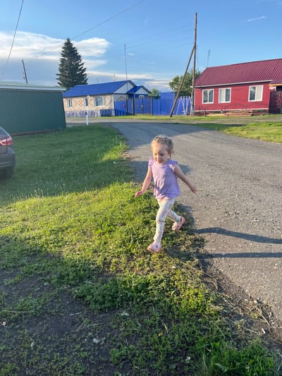 In Mal'tsevo, Russia, on a sunny day, a young blonde girl with pigtails adorned with watermelon-shaped clips runs on a gravel road. She is wearing a purple t-shirt with ruffled sleeves and patterned leggings, along with pink clogs. The girl is in motion, with her arms outstretched and her right leg forward as if mid-stride. To her left, a patch of green grass is illuminated by the sun, contrasting with the darker earth. Behind her, a road stretches towards the background, where a red-roofed house with white trim and windows stands prominently. Further left, a green building is visible, and beyond that, a house with a blue roof and white fence. A tall evergreen tree is situated near the blue-roofed house, and utility poles with wires run across the bright blue sky dotted with fluffy white clouds. The overall scene depicts a rural residential area on a clear day.