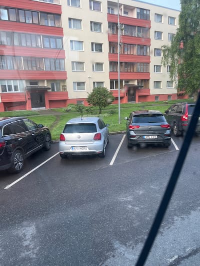 The image shows a parking lot in front of a residential apartment building in Maardu, Estonia. The building is tall and appears to be made of concrete with red and beige colored panels. There are several cars parked in the lot, including a black SUV, a silver Volkswagen Polo, a dark gray Volkswagen T-Roc, and a dark colored Volvo SUV. The ground is wet, indicating that it may have recently rained or is raining. There are also some trees and bushes visible in the background. The parking lot is marked with white lines to delineate the parking spaces. The overall scene is a typical residential parking area in an urban environment.