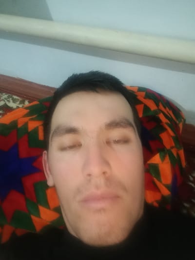 A close-up selfie depicts a man lying down, eyes nearly closed. He has short, dark hair and light skin. The background is a colorful patterned pillow or cushion, predominantly in shades of orange, green, and blue, with a brown wooden edge visible behind it. A white cylindrical object appears along the top of the frame, potentially a pipe or rod attached to the wall. The man is wearing a black shirt. The lighting is somewhat dim.