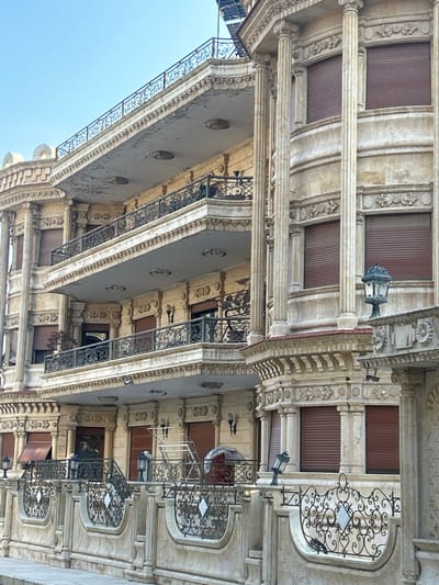This image, when viewed upright, displays the ornate stone facade of a multi-story building, likely situated in Aleppo, Syria, given the provided context. The building exhibits a classical or neoclassical architectural style with intricate detailing under a clear, bright blue sky, suggesting daytime.
The facade is constructed from light-colored stone or stucco, showing some signs of age but largely well-preserved. It features numerous decorative elements, including fluted columns, elaborate cornices, and detailed carvings such as floral motifs and scrolls, which adorn the lintels, pilasters, and sections between floors.
Multiple levels of the building are visible, each with windows or balcony doors, many of which are covered by brown, ribbed roll-down shutters. Projecting from the building are numerous balconies, each protected by elaborate black wrought-iron railings with intricate swirling patterns. Some lower-level openings appear to be recessed or framed by arches. Wall-mounted lanterns with dark metal fixtures and clear glass are affixed to the facade at various points, suggesting evening illumination.
No people or discernible activity are present, making the scene appear quiet and still. There is no visible, legible text on the building. The overall impression is one of historical grandeur and meticulous architectural craftsmanship.