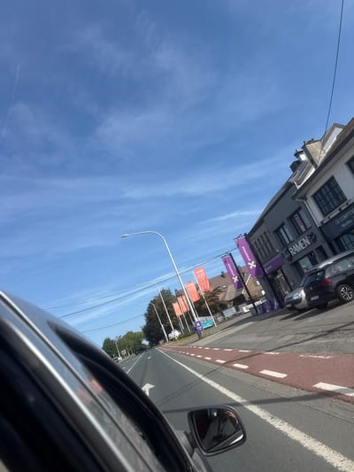 The image shows a view from inside a car looking towards the right, with the front of the car on the left side of the frame. It is a sunny day in Mont-Saint-Guibert, Belgium. The road is visible, with traffic lanes and a red bike lane. On the right side of the road, there are buildings with shops, including "RAMEN" and "LE COMPTOIR". Purple and orange flags are displayed outside some of the shops. A few cars are parked on the side of the road. The sky is blue with some wispy clouds and a contrail from an airplane.