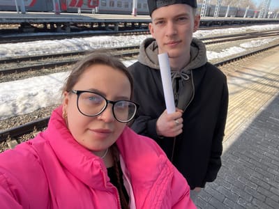 Two individuals stand close together, a man and a woman, in front of train tracks. The woman is wearing a pink jacket and glasses. The man wears a black jacket and a black baseball cap. They are at a train platform, with snow visible along the tracks. A train is in the background. The location is likely Tashevka, Russia. They are holding a white object, possibly a receipt or a ticket.