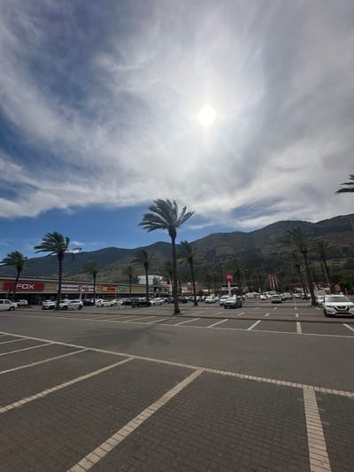 The image depicts a parking lot in the city of Galil Elyon Regional Council, Israel. Rows of cars are parked along the edge of the lot next to a building. Palm trees line the area between the parking lot and the building. A mountain is visible in the background behind the building. The sky is blue with scattered clouds, and the sun is shining brightly.