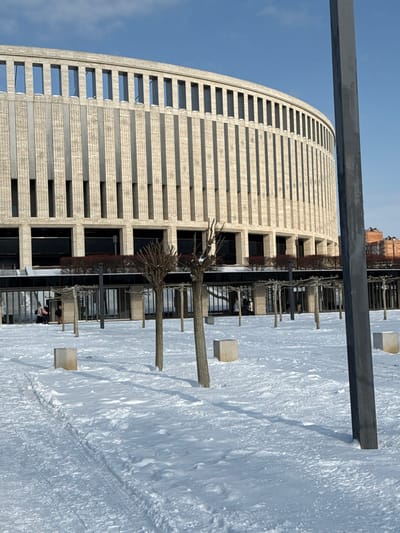 An outdoor scene depicts a snow-covered plaza in front of the Krasnodar Stadium, located in Plodorodny, Krasnodar, Russia, under a clear blue sky. The prominent subject is the stadium's large, curved, light-beige facade, characterized by numerous vertical textured panels and rectangular window-like openings across its upper and lower sections. Below the main facade, a darker, recessed ground level features multiple glass-fronted entrances and supporting pillars.
The foreground consists of a flat expanse of undisturbed snow, marked by visible footpaths and tire tracks. Scattered across this snow are several small, deciduous trees with pruned, bare branches, casting long shadows to the right. Also present are several light-colored, roughly cubic concrete blocks. To the far right, a tall, dark grey rectangular utility pole or mast stands vertically, partially obstructing the view of the stadium.
In the midground, near the stadium's lower level, several dark vertical posts support a dark, linear canopy structure. A few indistinct human figures, appearing as dark shapes against the building's lighter facade, are visible near the entrances on the left side, indicating foot traffic. The scene suggests a clear, cold winter day.
