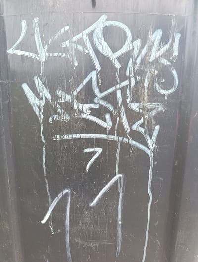 The image shows graffiti in white spray paint on a dark, weathered surface, likely a trash receptacle or utility box. The graffiti consists of abstract lettering and a number "7" followed by two vertical strokes that resemble the number "11." The spray paint is streaked and dripping in places, indicating it was applied in wet conditions or the surface is exposed to the elements. The background surface is scratched and dirty, with visible vertical lines suggesting it's a textured material. The scene is likely located outdoors in an urban or semi-urban environment. There are no people or specific landmarks visible that would pinpoint a location within Mechernich, Germany.