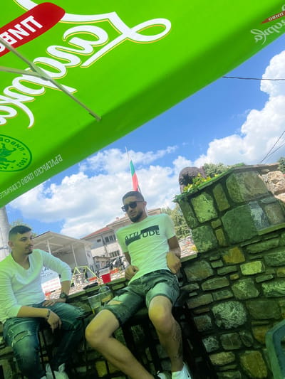 The image captures two men seated outdoors under a large green umbrella on a bright day with a blue sky and scattered white cumulus clouds, in Касапите, Bulgaria.

The green umbrella, prominent in the upper frame, displays white script lettering for "Каменица" (Kamenitza) and "UNBO," along with "БЪЛГАРСКО ПИВО С УКРЕПЛЕНИЕ" (Bulgarian Beer with Fortification) and "1881." A red stripe on the umbrella also shows "UNBO" in white.

Two men are seated on dark stools in the lower half of the frame, positioned in front of a stone wall.
The man on the left has short dark hair and stubble, dressed in a white long-sleeved shirt, distressed blue jeans, and white sneakers. He wears a watch and bracelet on his wrists and a ring on his left hand. He is looking towards the right of the frame, away from the camera. A smartphone, sunglasses, and a glass containing a clear liquid are on a table or ledge in front of him.
The man on the right has short dark hair and a beard, wearing dark sunglasses, a white short-sleeved t-shirt with "ATTITUDE" printed in black, distressed denim shorts, and white Nike Air Max 270 sneakers. A tattoo is visible on his right calf. He is looking directly at the camera, leaning against a stone wall structure. A glass with a dark liquid and a red plastic cup are present on the stone wall beside him.

A robust stone wall, constructed from varied-sized natural stones, extends across the midground. Green foliage and a dark, possibly bear-shaped, statue are positioned atop this wall.
In the background, a Bulgarian flag (horizontal green, white, and red stripes) is visible on a flagpole. Several buildings are present, including a white kiosk with legible text such as "ЕФИ 1994," phone numbers, "24/7," and "ТОПЛА ВОДА, ВАКСА" (Hot Water, Wax). Trees are visible beyond the buildings. A thin black electrical wire traverses the sky in the upper right portion of the image.