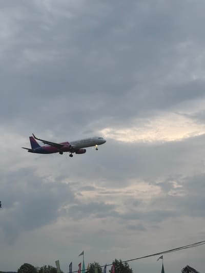 An airplane, identified as a Wizz Air aircraft by its distinctive purple/pink livery and visible text on the fuselage, is captured mid-flight against a cloudy sky. The plane appears to be either ascending shortly after takeoff or descending for landing, with its landing gear extended. The main body of the aircraft is white, while the tail and underside of the wings feature the characteristic purple/pink coloring.

The sky is predominantly overcast with varying shades of gray clouds, though a brighter, lighter patch of clouds is visible towards the upper right, suggesting the sun might be obscured behind them or it's late afternoon. In the background, to the right side of the frame, trees, utility lines, and a series of colorful decorative flags are partially visible, indicating a ground-level vantage point looking upwards. The overall lighting suggests an overcast day or possibly late afternoon. The setting is consistent with an airport vicinity, specifically near Otopeni, Romania, which hosts Henri Coandă International Airport (OTP).