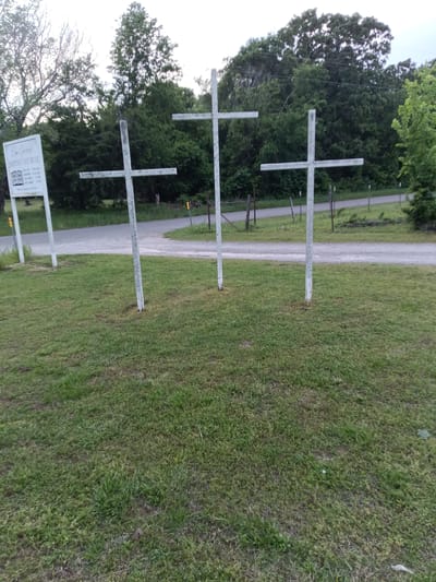 The image depicts three white crosses standing on a grassy lawn in front of a road. To the left is a sign for "Elm Grove Baptist Church." The background includes trees and foliage. The location is Stilwell, United States, based on the reference to a Baptist church in the area.