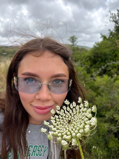 A young woman wearing translucent rimless sunglasses and a grey "Jersey" t-shirt is seen from a close-up, slightly angled perspective. She holds a cluster of white Queen Anne's Lace flowers near her face. The background consists of green foliage and an overcast sky. This photograph was likely taken outdoors, although the precise location in Nesher, Israel is impossible to ascertain from the limited visual data.