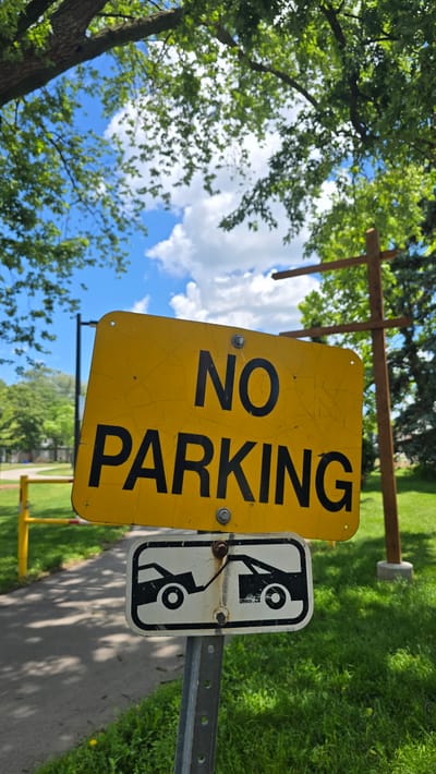 A yellow "NO PARKING" sign with a black tow-away symbol underneath is mounted on a metal pole. The "NO PARKING" sign is cracked. The tow-away symbol shows two cars, one being towed. The sign is located outdoors with green grass and trees in the background. A paved path is visible to the left of the sign, with a yellow barrier. The sky is blue with white clouds. The scene appears to be in a park or public area.