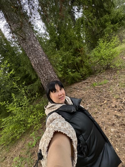 A medium shot depicts a person with dark hair and fair skin standing outdoors near a large tree in what appears to be a wooded area. The person is wearing a black puffer jacket and a tan patterned scarf. The ground is covered with low-lying greenery, interspersed with patches of dry, brown vegetation. The large tree is the focal point of the left side of the image, with its trunk and branches extending upwards into a dense canopy of green leaves and needles. The location is identified as Kamennoye, Russia. The person is looking at the camera.