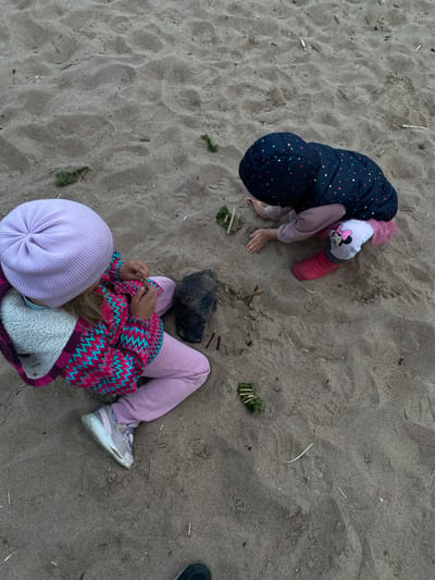 Two young children are playing in the sand on a beach in Baltiiskoe, Russia. The child on the left is wearing a lavender beanie, a patterned jacket, and pink pants. They are sitting with their legs crossed and are holding a small dark rock. The child on the right is wearing a hooded jacket with colorful stars and pink pants with a Minnie Mouse design. They are crouching down and interacting with small sticks and green debris in the sand. The environment is a sandy beach with scattered bits of driftwood and dried grass. The lighting suggests it may be overcast or late in the day.